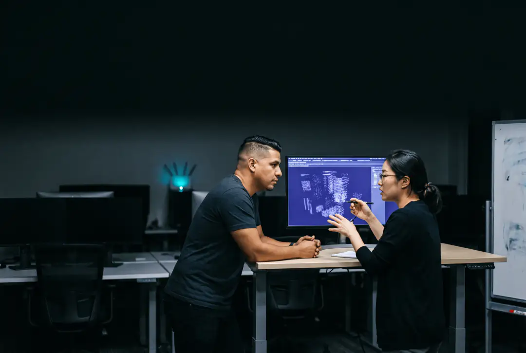 Two colleagues at a desk in a dark office, one pointing at a screen while the other explains a plan.